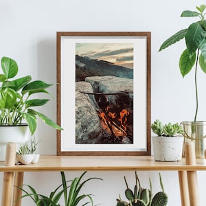 May include: A framed photograph of a campfire burning on a rocky cliff overlooking a sunset. The photo is displayed on a wooden shelf with plants and other decor.
