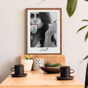 May include: Black and white photo of a woman holding a cup with a flower design. The photo is framed in a brown frame and has the text "قَوِيَّة" written below the image.