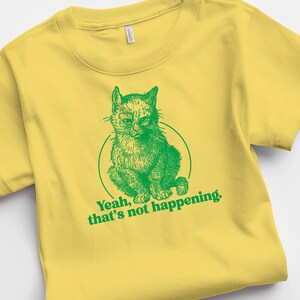 Op de afbeelding: Geel T-shirt met een groene illustratie van een kat met de tekst "Yeah, that's not happening."