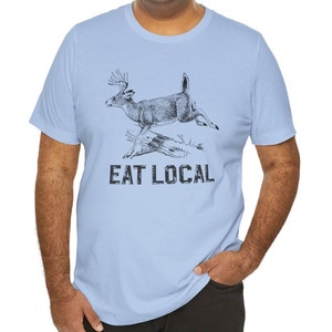 May include: Light blue t-shirt with a black and white illustration of a deer jumping over a log. The text "EAT LOCAL" is printed below the deer.