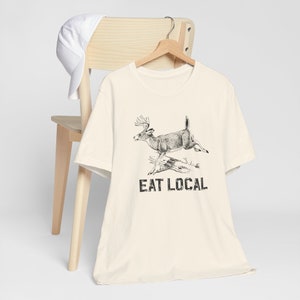 May include: A cream-colored t-shirt with a black and white illustration of a deer jumping over a log. The text "EAT LOCAL" is printed below the deer.