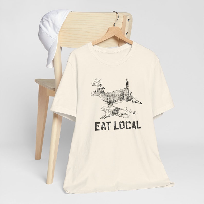 May include: A cream-colored t-shirt with a black and white illustration of a deer jumping over a log. The text "EAT LOCAL" is printed below the deer.