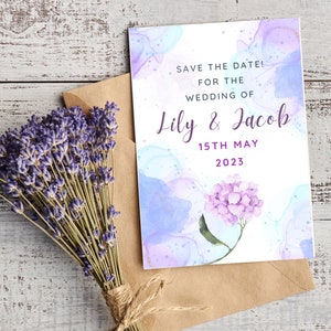 Puede incluir: Tarjeta de guardar la fecha con fondo de acuarela en tonos azules y morados. La tarjeta presenta el texto "Save the Date! For the Wedding of Lily & Jacob 15th May 2023" y una flor de hortensia morada.