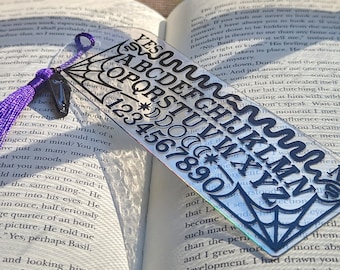May include: A clear acrylic bookmark with black lettering and numbers. The design includes the alphabet, numbers, and the word "YES". A purple tassel and a black charm are attached to the top. The bookmark is placed in an open book.