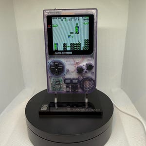 Puede incluir: Una consola portátil Game Boy Color de color púrpura transparente que muestra un videojuego clásico. La pantalla muestra un nivel del juego. La consola está montada en un soporte giratorio negro, con las palabras "GAME BOY COLOR" visibles.