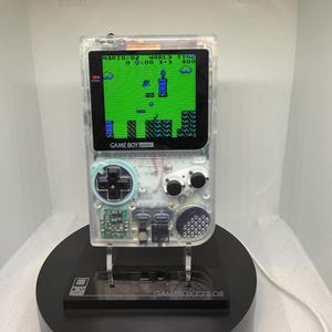 Puede incluir: Una consola portátil Game Boy Pocket transparente, que muestra un nivel del juego Mario 02. La consola está montada en un soporte de exhibición negro con las palabras "GAMEBOY COLOR" visibles. La pantalla muestra un fondo verde con gráficos pixelados azules y negros.