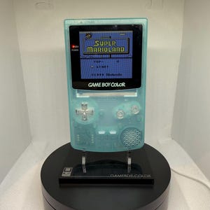 Könnte beinhalten: Ein transparentes, türkisblaues Game Boy Color Handheld-Konsolen-Gerät, das das Spiel "Super Marioland" anzeigt. Der Bildschirm zeigt den Titel des Spiels und das Startmenü. Die Konsole steht auf einem schwarzen Ständer mit der Aufschrift "GAME BOY COLOR".