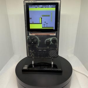 May include: A transparent Game Boy Color handheld console displaying a level from Super Mario Bros. The console is mounted on a black display stand. The screen shows a pixelated landscape with a character and obstacles.