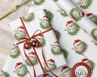 May include: White wrapping paper featuring a repeating pattern of a cat wearing a Santa hat and a wreath. A wrapped gift is tied with red ribbon and adorned with red ornaments. Gold scissors are visible.