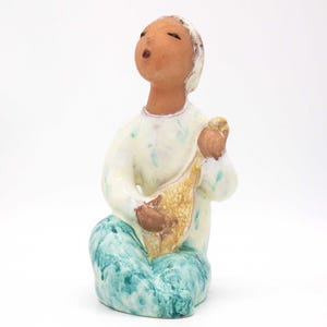 Könnte beinhalten: Eine Keramikfigur einer sitzenden Person mit einem Musikinstrument. Die Person trägt ein weißes Hemd mit grün-weißen Akzenten und eine blaue Hose. Das Instrument ist braun und gelb.