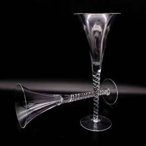 Könnte beinhalten: Zwei klare Champagnerflöten aus Glas mit einem gedrehten Stiel. Die Flöten stehen auf einem schwarzen Hintergrund.