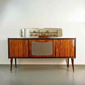 Könnte beinhalten: Ein Sideboard im Stil der Moderne aus der Mitte des Jahrhunderts mit einer Holzfurnier-Oberfläche und Messingbeschlägen. Das Sideboard hat drei Türen, von denen eine ein Glaspaneel mit einem dekorativen Design aufweist. Das Sideboard wird von vier schlanken Beinen getragen.
