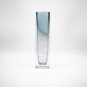 Könnte beinhalten: Eine klare Glasvase mit einer strukturierten Oberfläche und einem hellblauen Farbton. Die Vase ist rechteckig und hat einen quadratischen Boden.