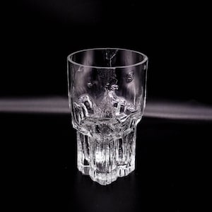 Könnte beinhalten: Ein klares, strukturiertes Glas mit einem einzigartigen Design. Das Glas hat ein detailliertes, geprägtes Muster auf der unteren Hälfte, mit einer zylindrischen Form und einer breiten, offenen Oberseite. Der Boden hat eine sternartige Form.