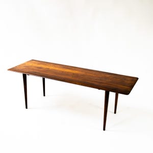 Puede incluir: Una mesa de centro rectangular de estilo moderno de mediados de siglo con un rico acabado de vetas de madera oscura. La mesa tiene cuatro patas cónicas y un diseño simple y aerodinámico. La mesa mide unos 152 cm de largo.