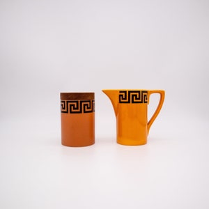 Può includere: Un set di stoviglie da cucina in ceramica arancione con un motivo geometrico nero. Il set comprende un bricco per la panna e una zuccheriera.