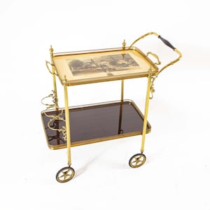 Op de afbeelding: Een vintage messing barwagen met twee rechthoekige planken. De bovenste plank heeft een ingelijste print, terwijl de onderste plank donkerbruin glas is. De wagen heeft decoratieve accenten, een handvat met een donkerhouten grip en kleine wielen.