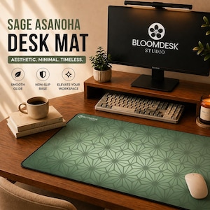 Op de afbeelding: Saliegroene bureaumat met een geometrisch patroon, een computermonitor, toetsenbord en muis op een houten bureau. De bureaumat heeft de tekst "Sage Asanoha Desk Mat" en "Aesthetic. Minimal. Timeless." erop gedrukt. Een koffiemok en boeken staan ook op het bureau.