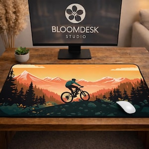 Può includere: Tappetino per mouse rettangolare con una scena di mountain bike al tramonto. Un ciclista attraversa una foresta, con un bordo nero. Un mouse bianco è accanto. Il monitor mostra il testo "BLOOMDESK STUDIO".