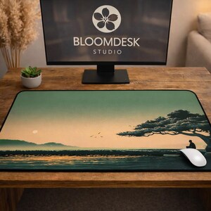 Puede incluir: Alfombrilla de ratón rectangular con un diseño de paisaje sereno que presenta un árbol, una persona y una escena oceánica. La alfombrilla está sobre un escritorio de madera con un monitor de computadora que muestra "BLOOMDESK STUDIO". Un ratón blanco está sobre la alfombrilla.