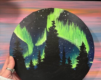 May include: A round wooden painting depicting a dark forest with a green aurora borealis in the sky. The painting is set against a pink and blue background.