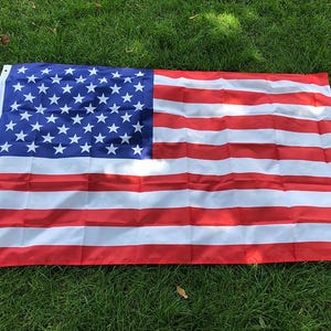 Puede incluir: Una bandera estadounidense rectangular con un cantón azul lleno de estrellas blancas y rayas rojas y blancas alternas. La bandera se muestra sobre una superficie de hierba verde.