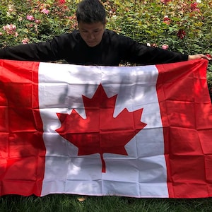 Puede incluir: Una bandera canadiense, con una hoja de arce roja sobre un cuadrado blanco, flanqueada por franjas verticales rojas. La bandera se muestra al aire libre, sostenida por una persona con una camisa negra de manga larga, sobre un fondo de hierba verde y flores.
