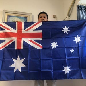 Puede incluir: Una bandera australiana azul con la Union Jack en la esquina superior izquierda y estrellas blancas. La bandera es rectangular y tiene un fondo azul oscuro. La bandera está siendo sostenida por una persona.