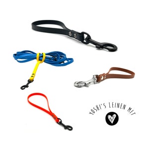 Op de afbeelding: Vier verschillende hondenriemen in verschillende kleuren, waaronder zwart, blauw, bruin en oranje. De riemen zijn gemaakt van een rubberachtig materiaal en hebben metalen clips. De tekst "YOSHI'S LEINEN MIT" is zichtbaar in de afbeelding.