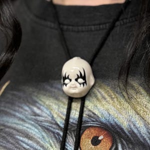 Peut inclure: Un pendentif en forme de tête de poupée en céramique blanche avec des détails noirs est suspendu à un collier en cordon noir. Le pendentif est porté sur un t-shirt noir avec un motif d'aigle détaillé.