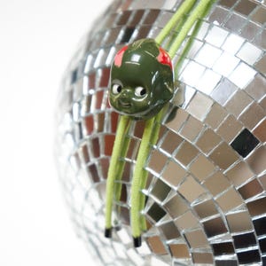 Peut inclure: Une boule disco argentée avec un visage en céramique verte. Le visage a des détails rouges et des cordons verts. La boule est recouverte de petits carrés réfléchissants.