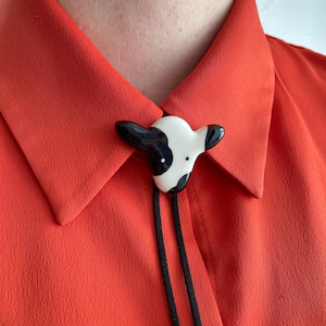 May include: A black and white cow-shaped bolo tie clasp with a black cord, set against a bright orange collared shirt. The clasp is ceramic and features a detailed cow face.