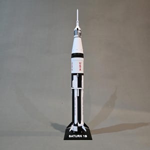 May include: A white and black model of a Saturn 1B rocket with a red "USA" inscription on the side. The rocket is on a black base with the words "Saturn 1B" printed on it.