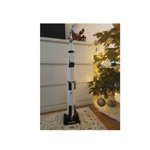 May include: A white and black model of the Saturn V rocket on a black base with the text "SATURN V" printed on it. The rocket is standing on a beige rug in front of a Christmas tree.