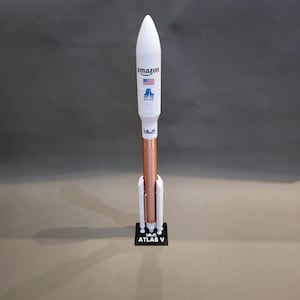 Könnte beinhalten: Ein Modellrakete mit weißer Spitze und kupferfarbenem Körper. Die Rakete trägt das Amazon-Logo und die amerikanische Flagge. Sie steht auf einem schwarzen Sockel mit der Aufschrift "ATLAS V".