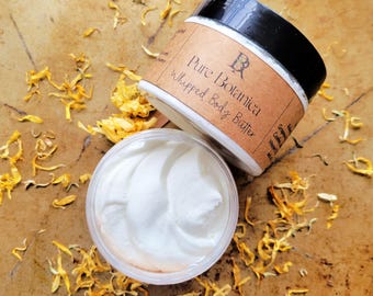 May include: Two jars of Pure Botanica Whipped Body Butter. One jar is open, revealing the white, whipped cream-like product. The other jar is partially visible, with a brown label and black lid. Dried yellow flower petals are scattered around the jars on a brown surface.