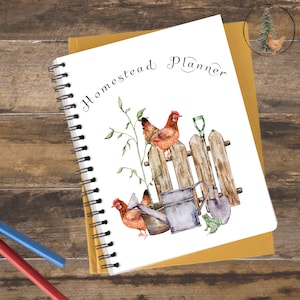 K&ouml;nnte beinhalten: Ein wei&szlig;es Spiralnotizbuch mit dem Text "Homestead Planner" in brauner Kursivschrift. Der Umschlag zeigt eine Aquarell-Illustration von zwei H&uuml;hnern, die auf einem Holzzaun sitzen, mit einer Gie&szlig;kanne, einer Schaufel und einem Frosch.