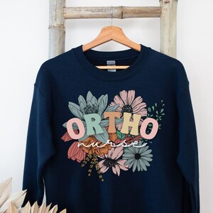 Peut inclure: Sweat-shirt bleu marine avec un motif floral et le texte "ORTHO nurse" en lettres color&eacute;es.
