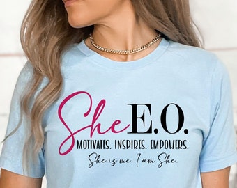 May include: A light blue t-shirt with the text "She E.O. MOTIVATES. INSPIRES. EMPOWERS. She is me. I am She." printed in black and pink.