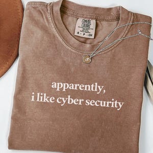 May include: Espresso-coloured t-shirt with the text "apparently, i like cyber security" in white. The shirt has a crew neck and a Comfort Colors tag. A silver necklace with a pendant is draped across the shirt.