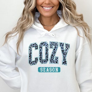 May include: White hooded sweatshirt with a blue and black leopard print graphic that says "Cozy Season".