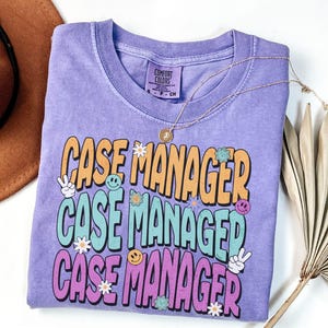 May include: Violet-colored t-shirt with the words "CASE MANAGER" in orange, teal, and purple, with peace signs, smiley faces, and flower graphics. A gold necklace with a pendant rests on the shirt.