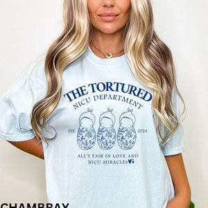 May include: Light blue t-shirt with a graphic design of three baby dolls with floral patterns. The text "The Tortured NICU Department Est. 2024 All's Fair in Love and NICU Miracles" is printed on the shirt.