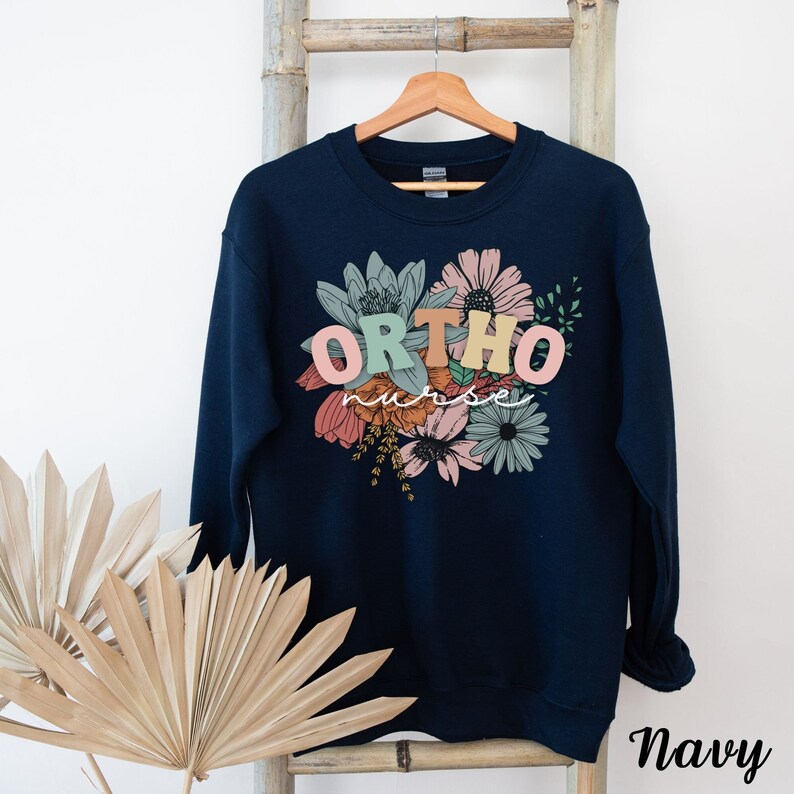 Peut inclure: Sweat-shirt bleu marine avec un motif floral et le texte "ORTHO nurse" en lettres color&eacute;es.
