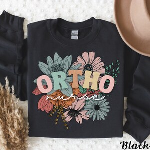 Peut inclure: Sweat-shirt noir avec un motif floral et le texte "ORTHO nurse" en lettres color&eacute;es.