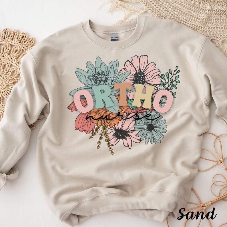 Peut inclure: Un sweat-shirt couleur sable avec un motif floral et le texte "ORTHO nurse" en lettres color&eacute;es.