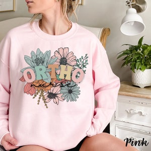 Peut inclure: Sweat-shirt rose avec un motif floral et le texte "ORTHO nurse" en lettres color&eacute;es.