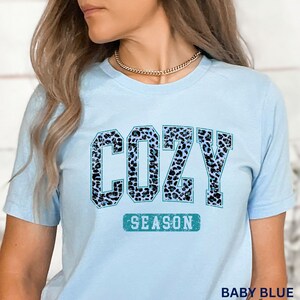 May include: A light blue t-shirt with a blue and black leopard print graphic that says "COZY SEASON".
