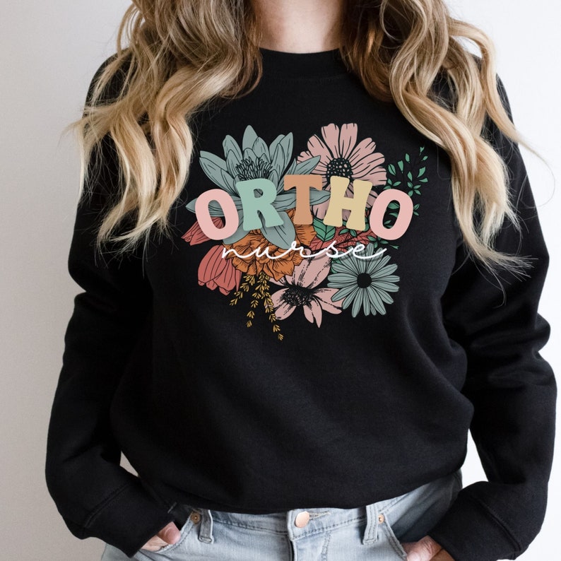 Peut inclure: Sweat-shirt noir avec un motif floral et le texte "ORTHO nurse" en lettres color&eacute;es.