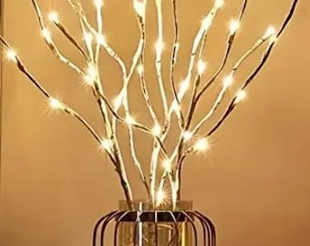 May include: A decorative arrangement featuring illuminated branches in a glass vase with a metal cage. The branches are adorned with small, warm white lights, creating a soft glow. A lit candle sits nearby.